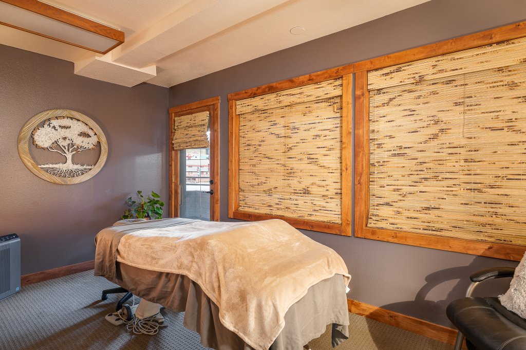 A serene massage therapy room with a massage table covered in a soft blanket, surrounded by wooden-framed bamboo blinds and adorned with a tree-themed wall art piece in Grand County Colorado.