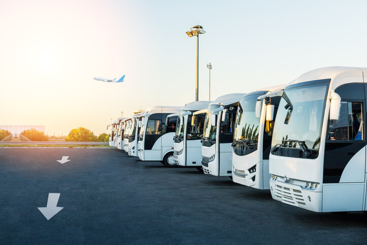 A row of white shuttle buses parked on an airport lot at sunrise, with an airplane taking off in the background.
