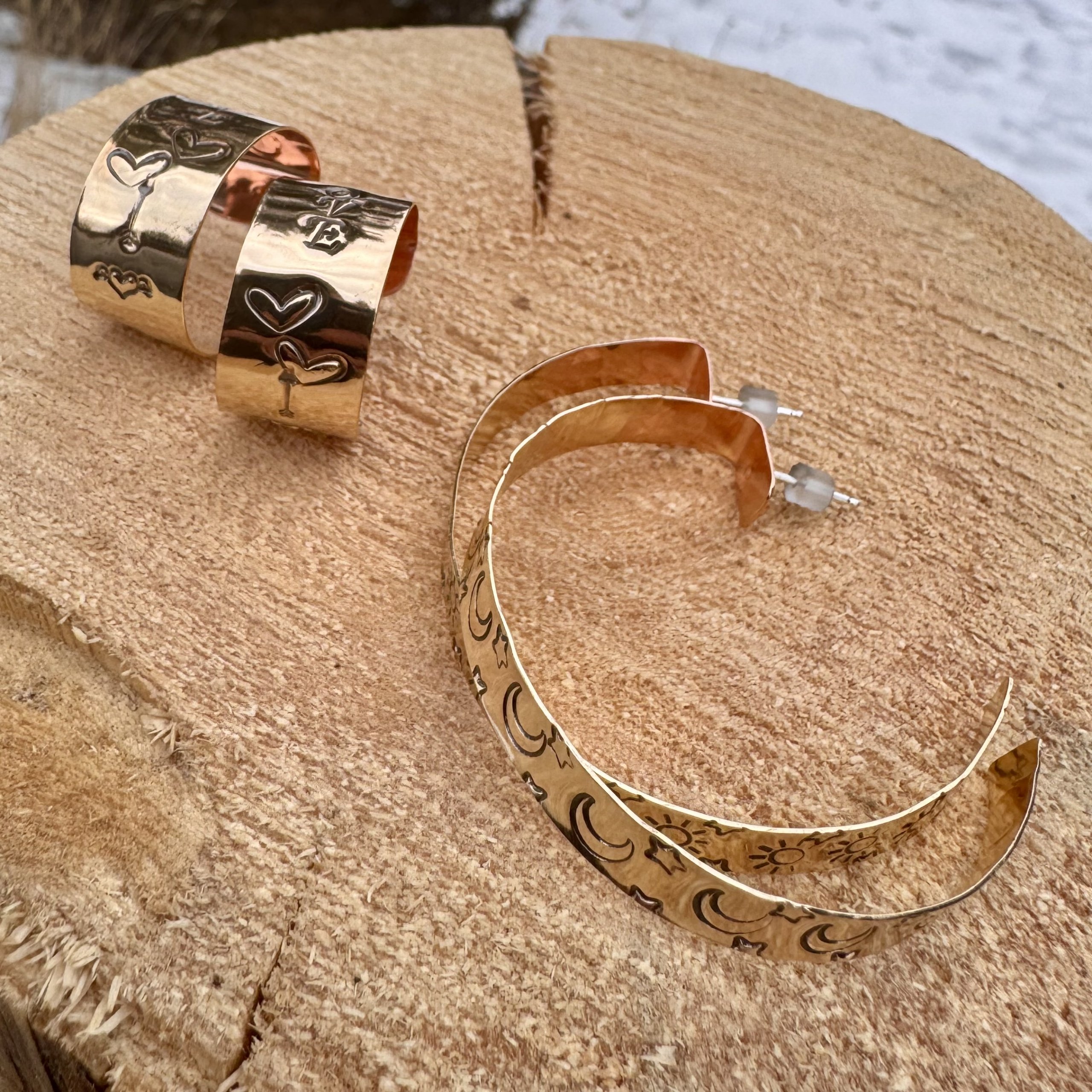 Three pairs of handmade hammered copper and gold hoop earrings with heart, star, and moon designs rest on a rustic wooden surface in Grand County Colorado.