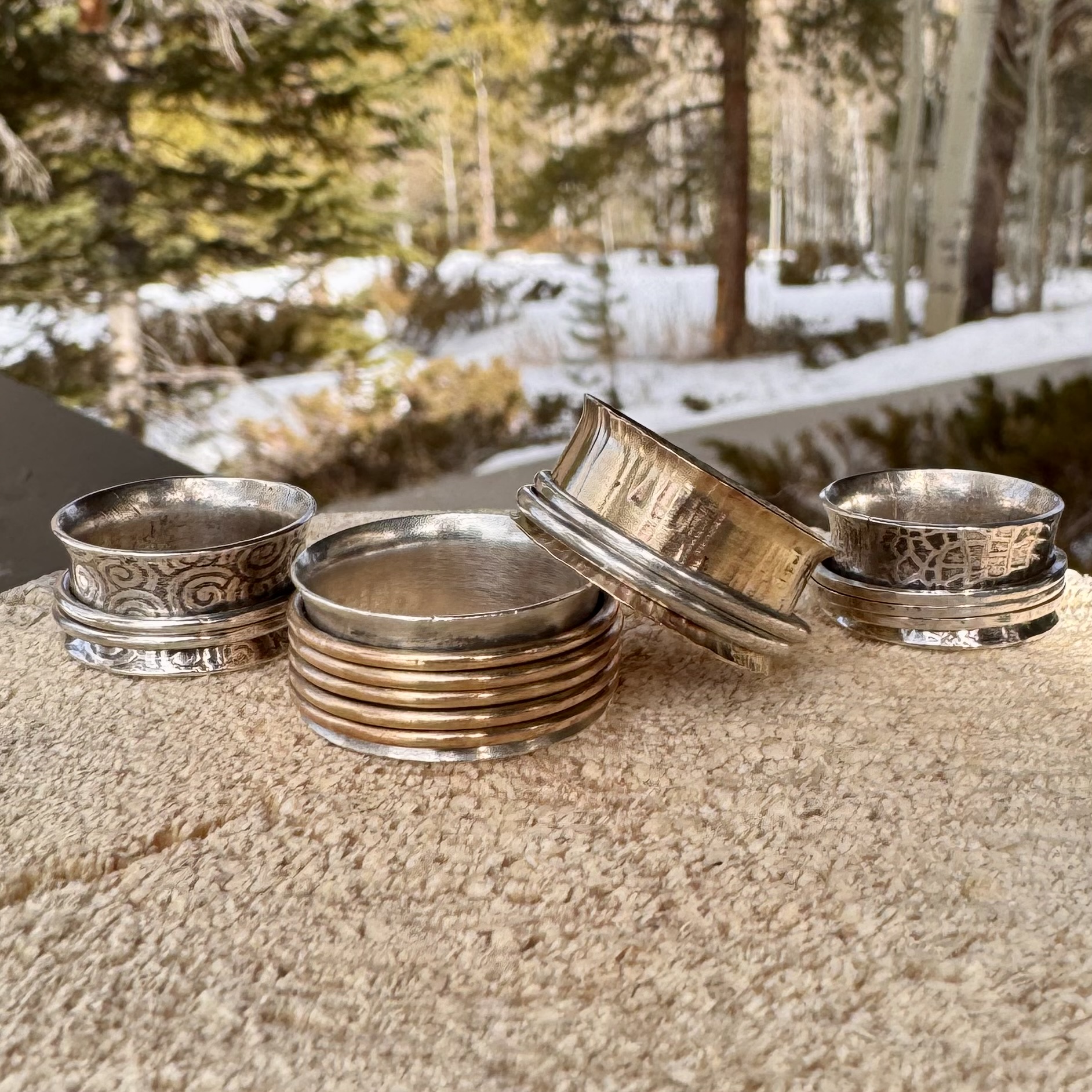 A collection of intricately designed silver and gold-toned spinning rings and bowls rests on a wooden surface, with a serene winter forest landscape in the background in Grand County Colorado.