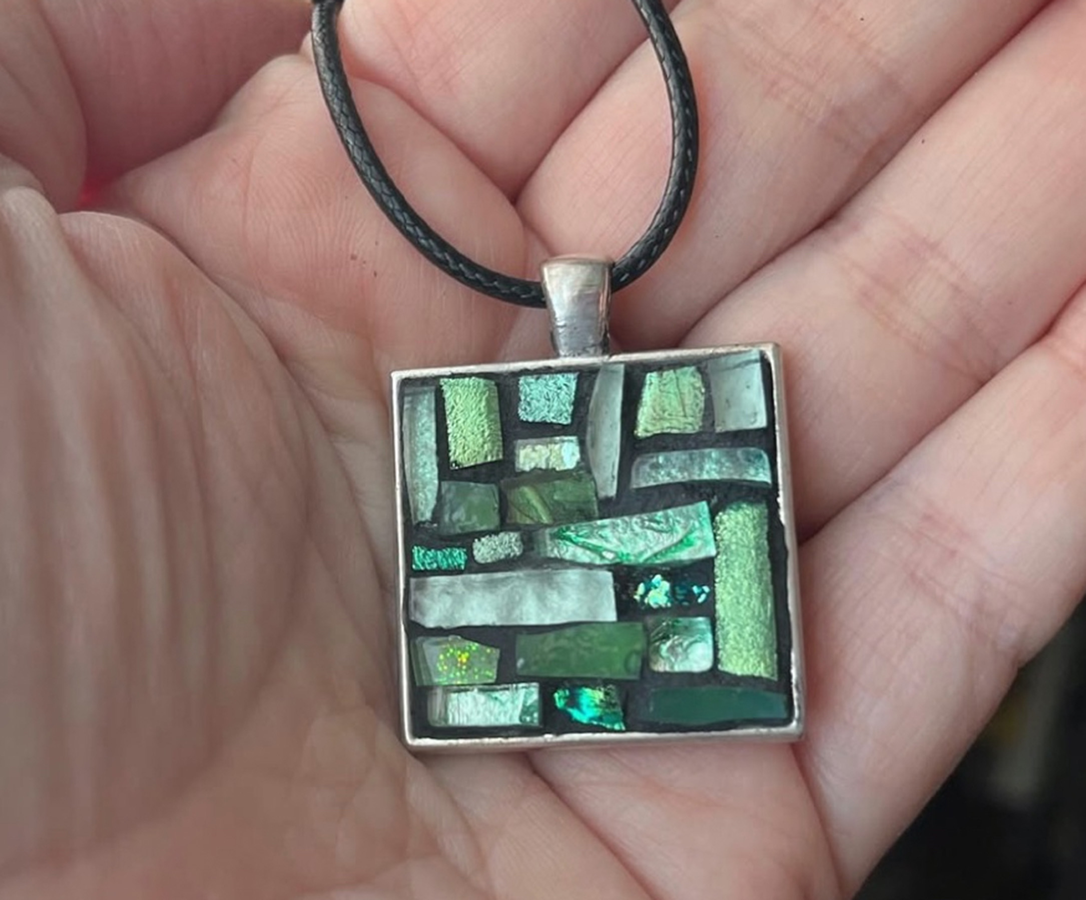 A person holding a square pendant with a mosaic of green and blue glass tiles in Grand County Colorado.