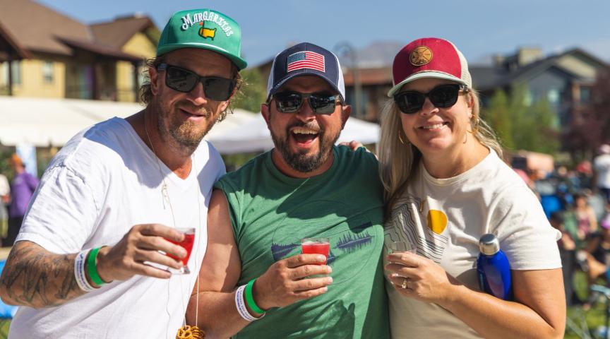 Three friends enjoying drinks and laughter at an outdoor event, wearing colorful hats and sunglasses in Grand County Colorado.