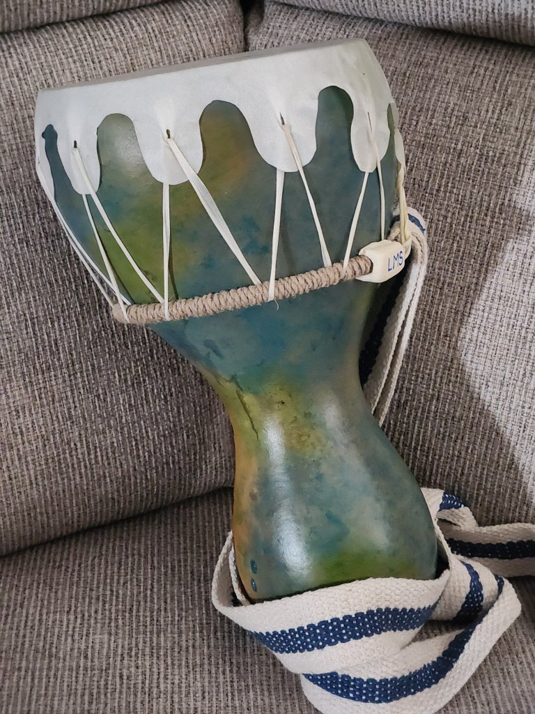 A hand-painted, hourglass-shaped drum with a white, wave-like rim and blue-green body rests on a gray couch, adorned with a white and blue striped strap in Grand County Colorado.