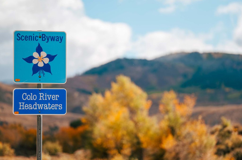 A close-up of the Colorado River Headwaters Scenic and Historic Byway road sign, and in the background, mountains and trees with fall colors.
