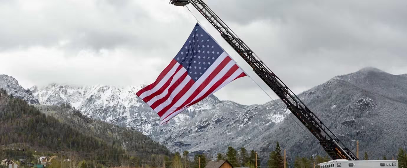 A fire truck ladder raises an American flag against a backdrop of snow-capped mountains and a cloudy sky in Grand County Colorado.