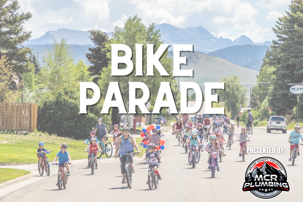 A colorful bike parade with children and adults riding bicycles down a suburban street, surrounded by trees and mountains in the background in Grand County Colorado.
