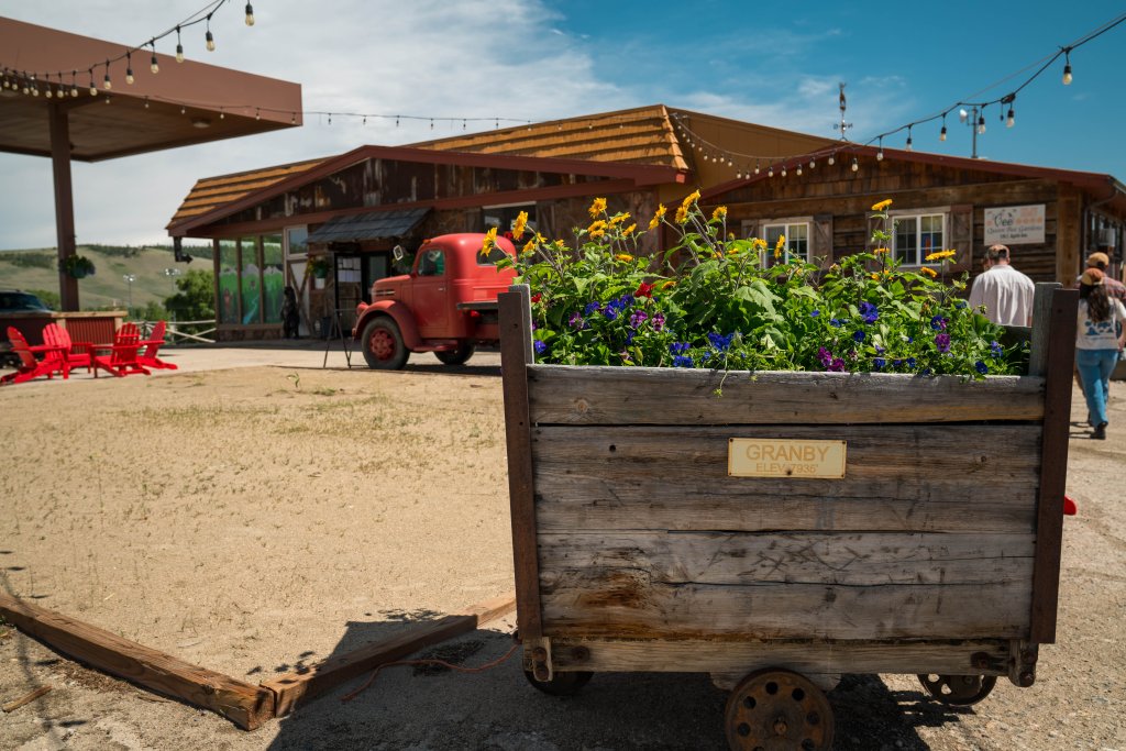 A rustic wooden flower cart filled with vibrant sunflowers and other colorful blooms stands in front of a charming, old-fashioned gas station with a red vintage truck parked nearby in Grand County Colorado.