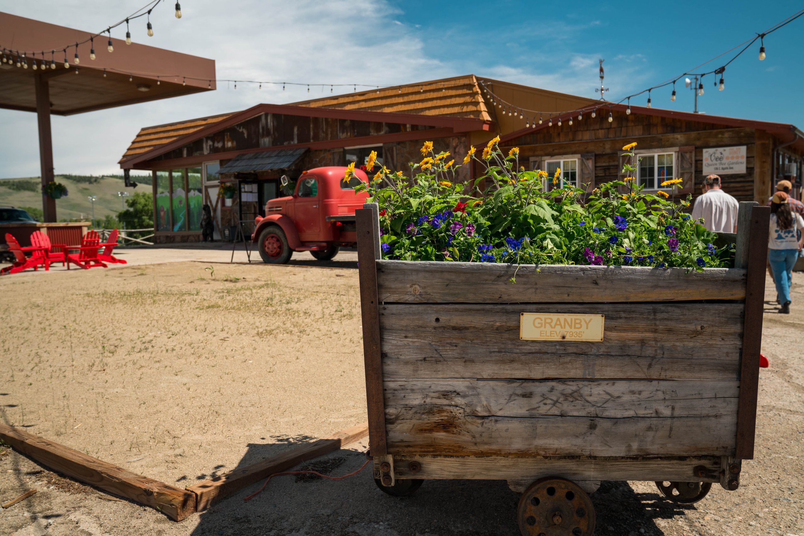 A rustic wooden flower cart filled with vibrant sunflowers and other colorful blooms stands in front of a charming, old-fashioned gas station with a red vintage truck parked nearby in Grand County Colorado.