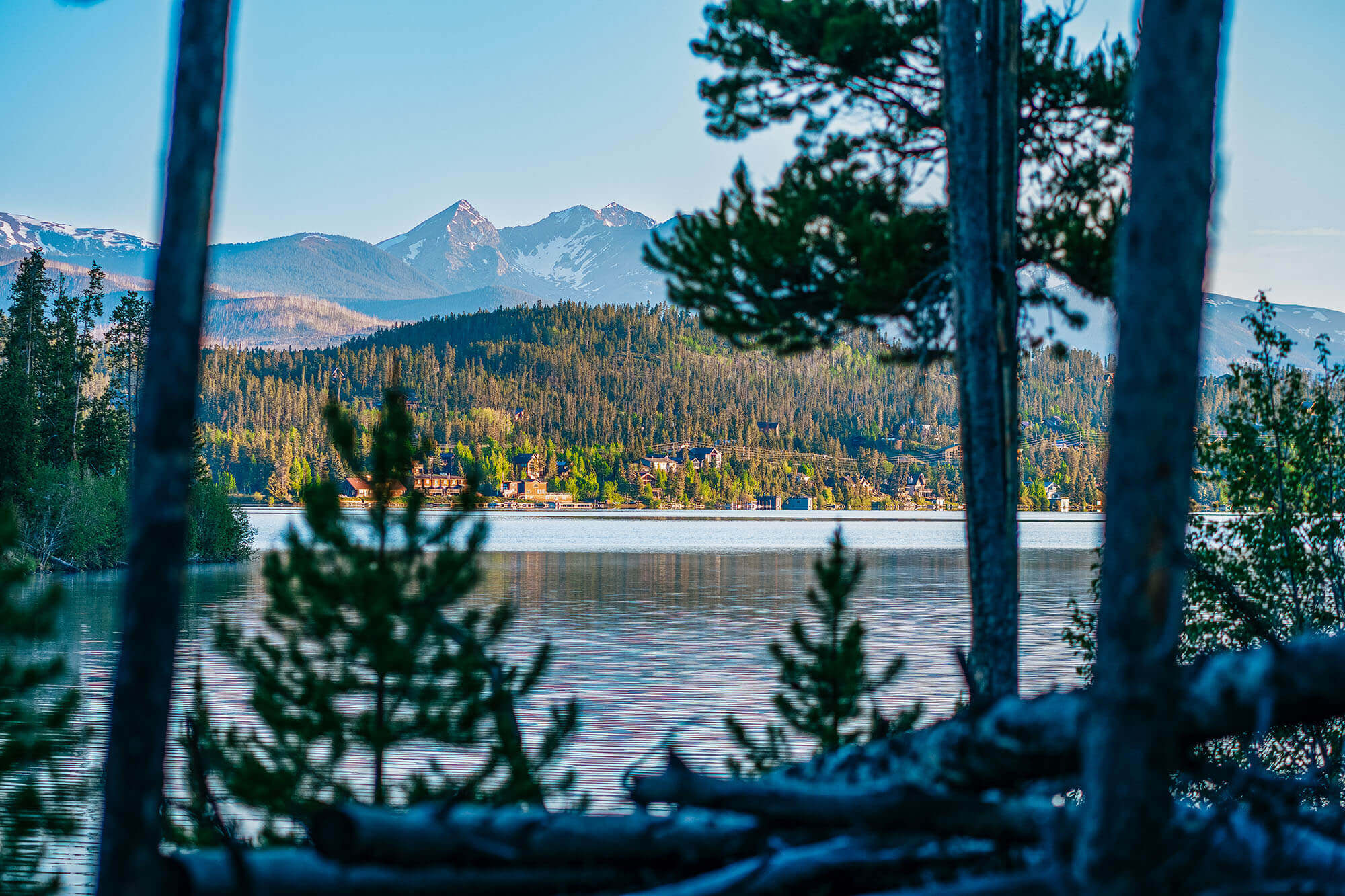 A serene lake nestled among towering mountains with snow-capped peaks, framed by lush green forests and tall trees in Grand County Colorado.