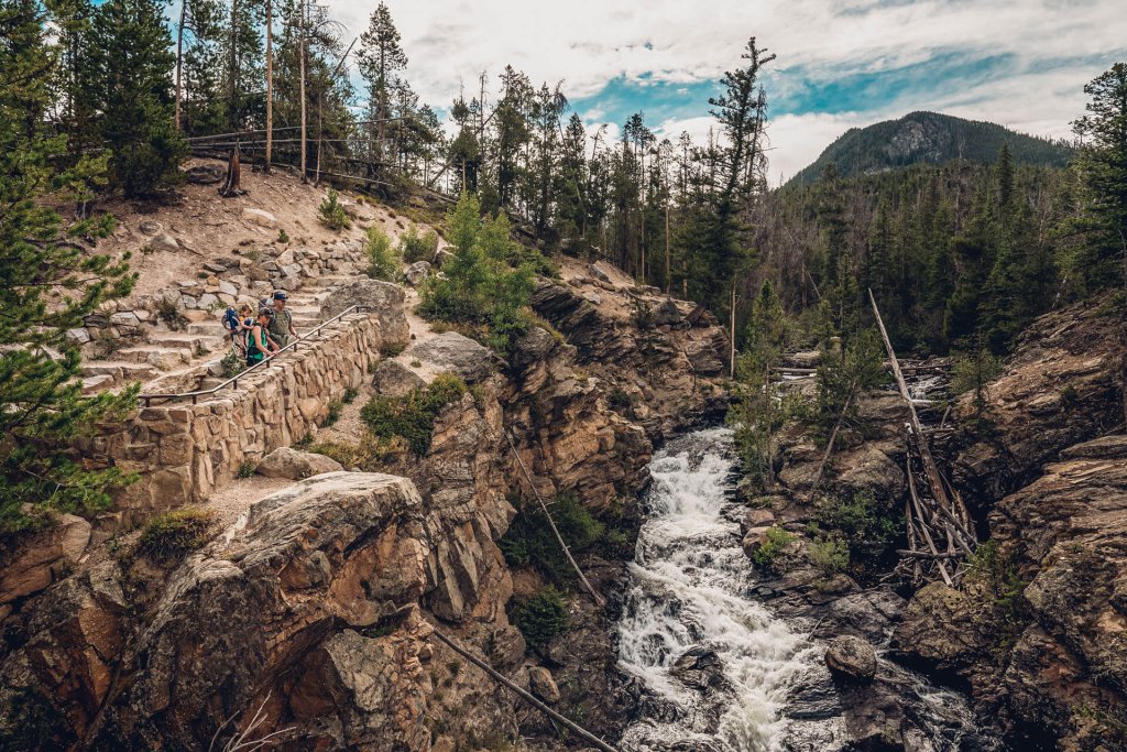 Hikers ascend a stone staircase along a rugged mountain trail, with a cascading waterfall and dense forest in the background in Grand County Colorado.