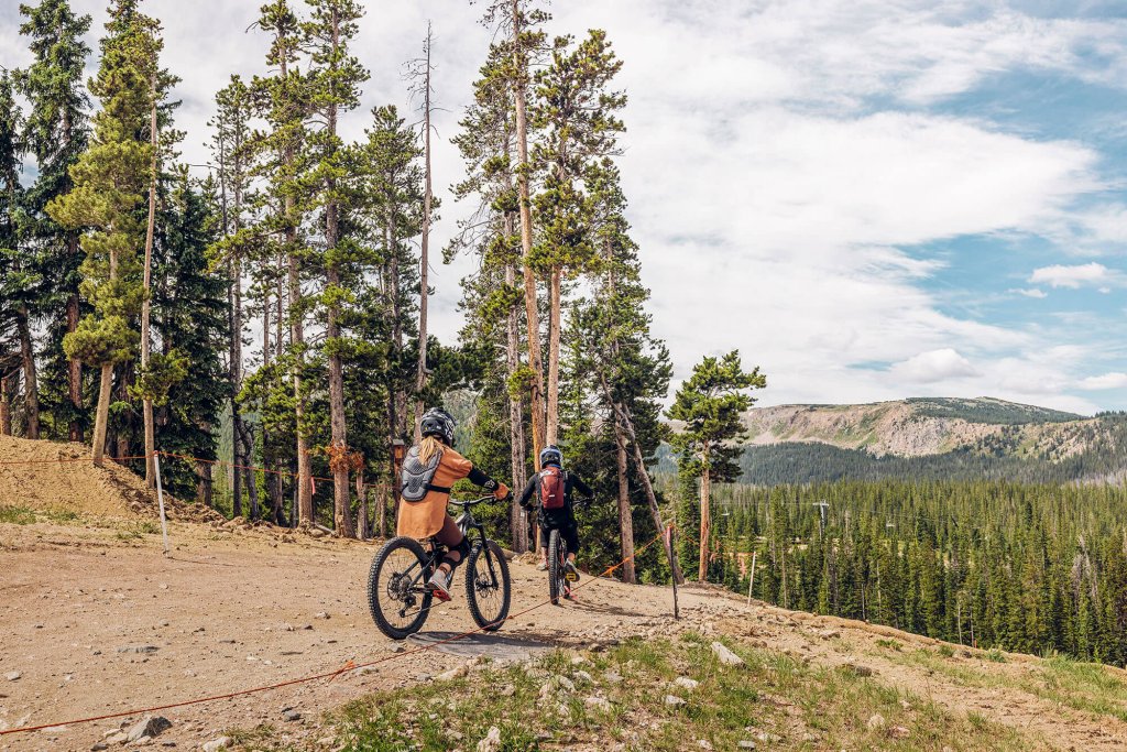 Two mountain bikers ride along a dirt trail through a pine forest with a scenic mountain view in the background in Grand County Colorado.