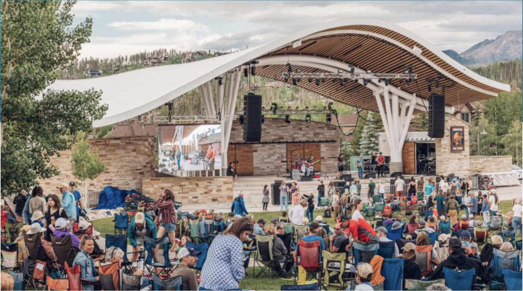 A large crowd enjoys an outdoor concert under a curved wooden pavilion with a mountain backdrop in Grand County Colorado.