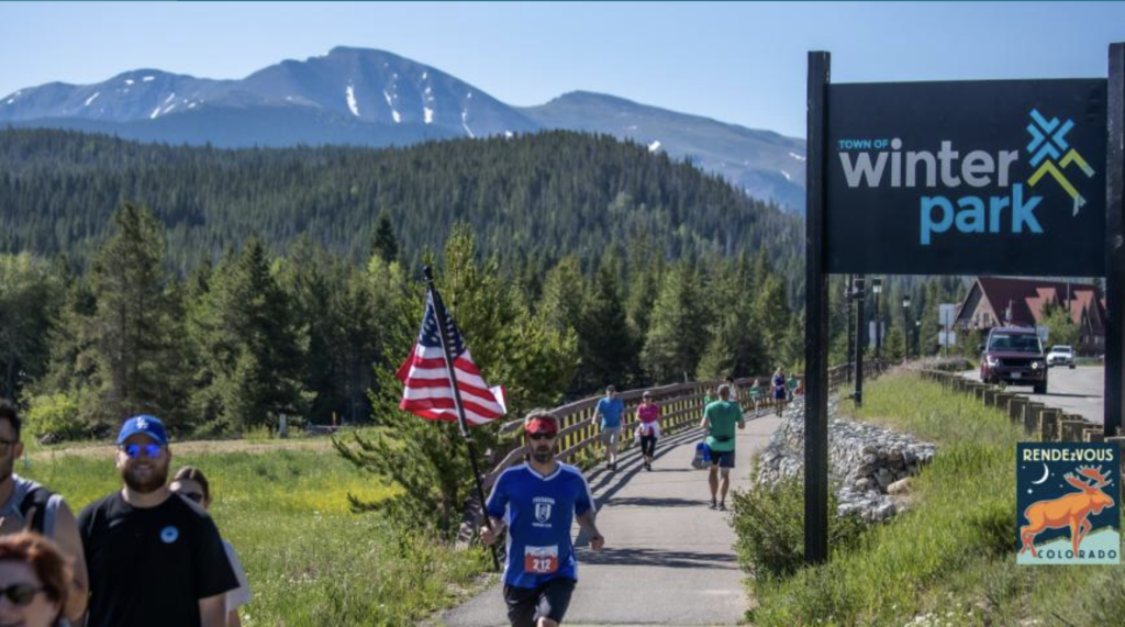 A group of runners participate in a race through Winter Park, Colorado, with a backdrop of snow-capped mountains and lush green forests in Grand County Colorado.