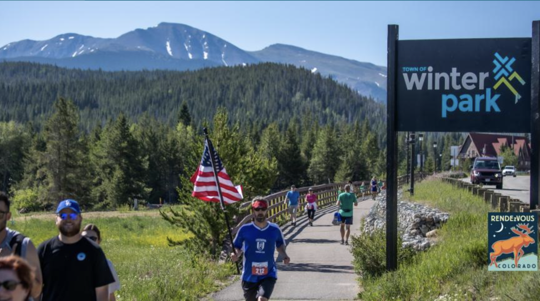 A group of runners participate in a race through Winter Park, Colorado, with a backdrop of snow-capped mountains and lush green forests in Grand County Colorado.
