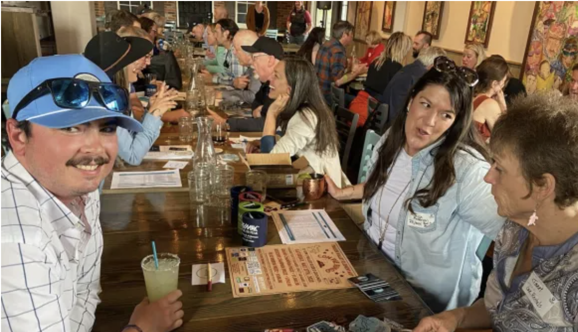 A lively group of people enjoying drinks and conversation at a long wooden table in a colorful, bustling bar in Grand County Colorado.