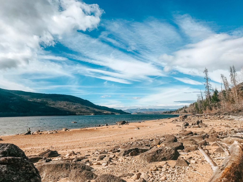 The sandy, rocky shores along Lake Granby in the foreground, and in the background, trees and mountains.