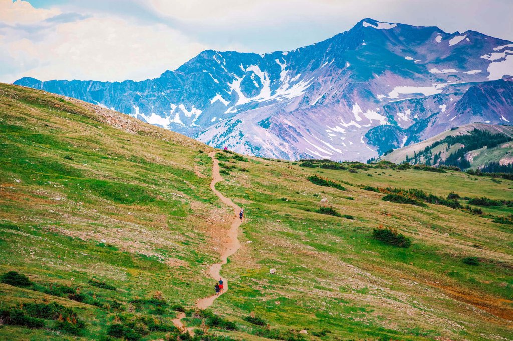A winding dirt path ascends a grassy mountain slope with distant snow-capped peaks in the background in Grand County Colorado.