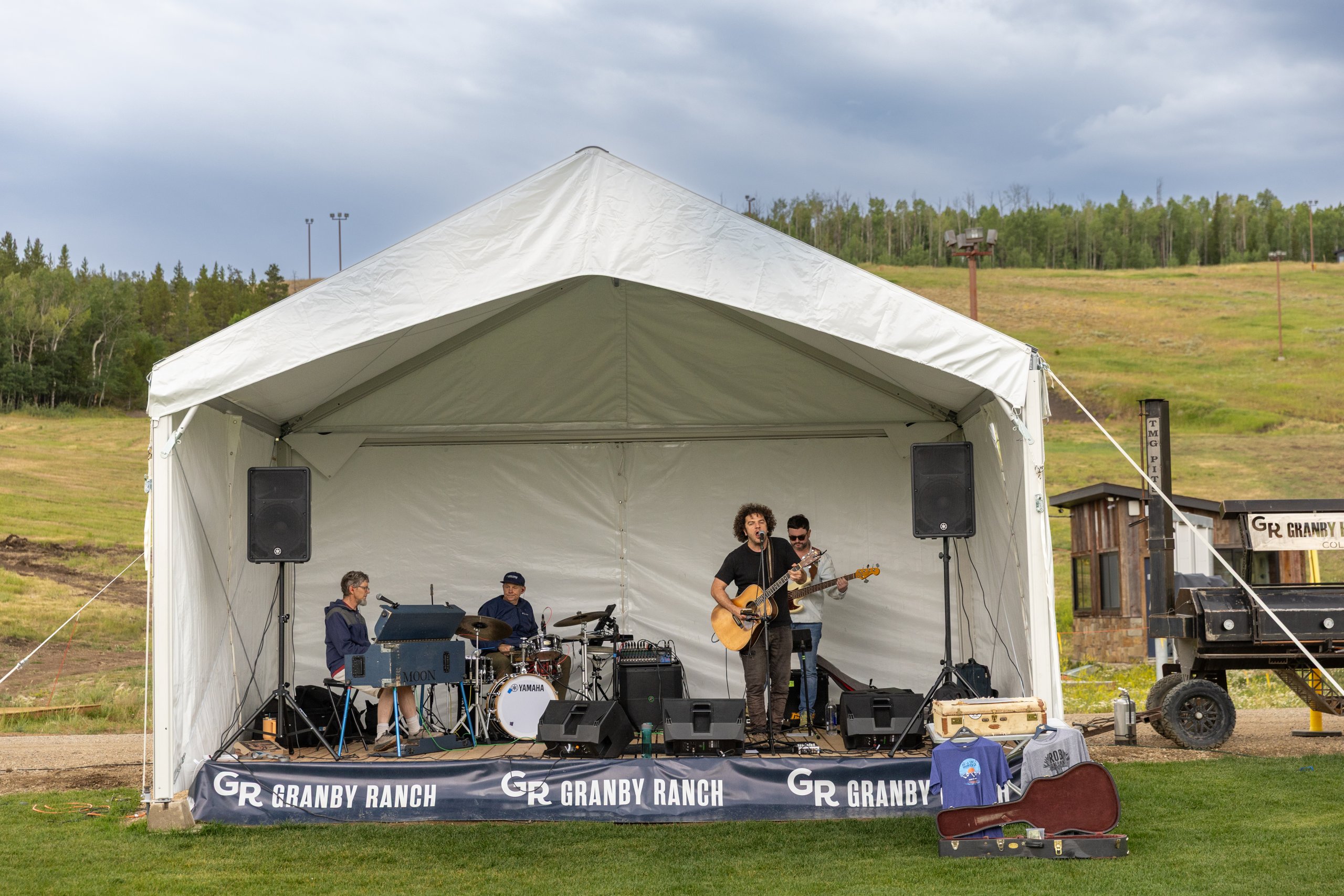 A band performs on a stage at Granby Ranch, with a white tent and speakers set up on a grassy field in Grand County Colorado.