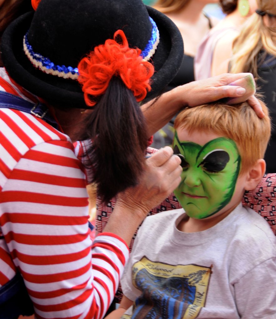 A clown wearing a black hat with a red flower and blue, white, and red trim is painting a green alien face on a young boy's face in Grand County Colorado.
