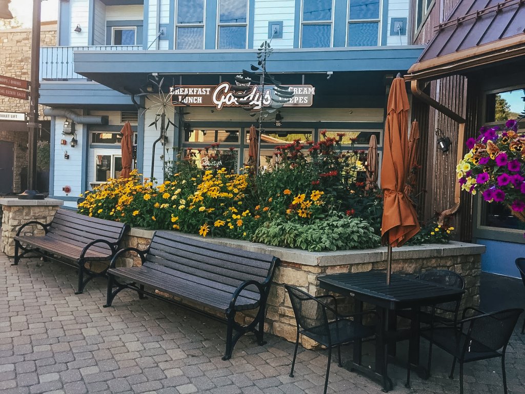 An outdoor seating area with wooden benches and tables surrounded by vibrant yellow and purple flowers, located in front of a restaurant with a blue exterior and a sign reading 'Goodys Mountain Creperie' in Grand County, Colorado.