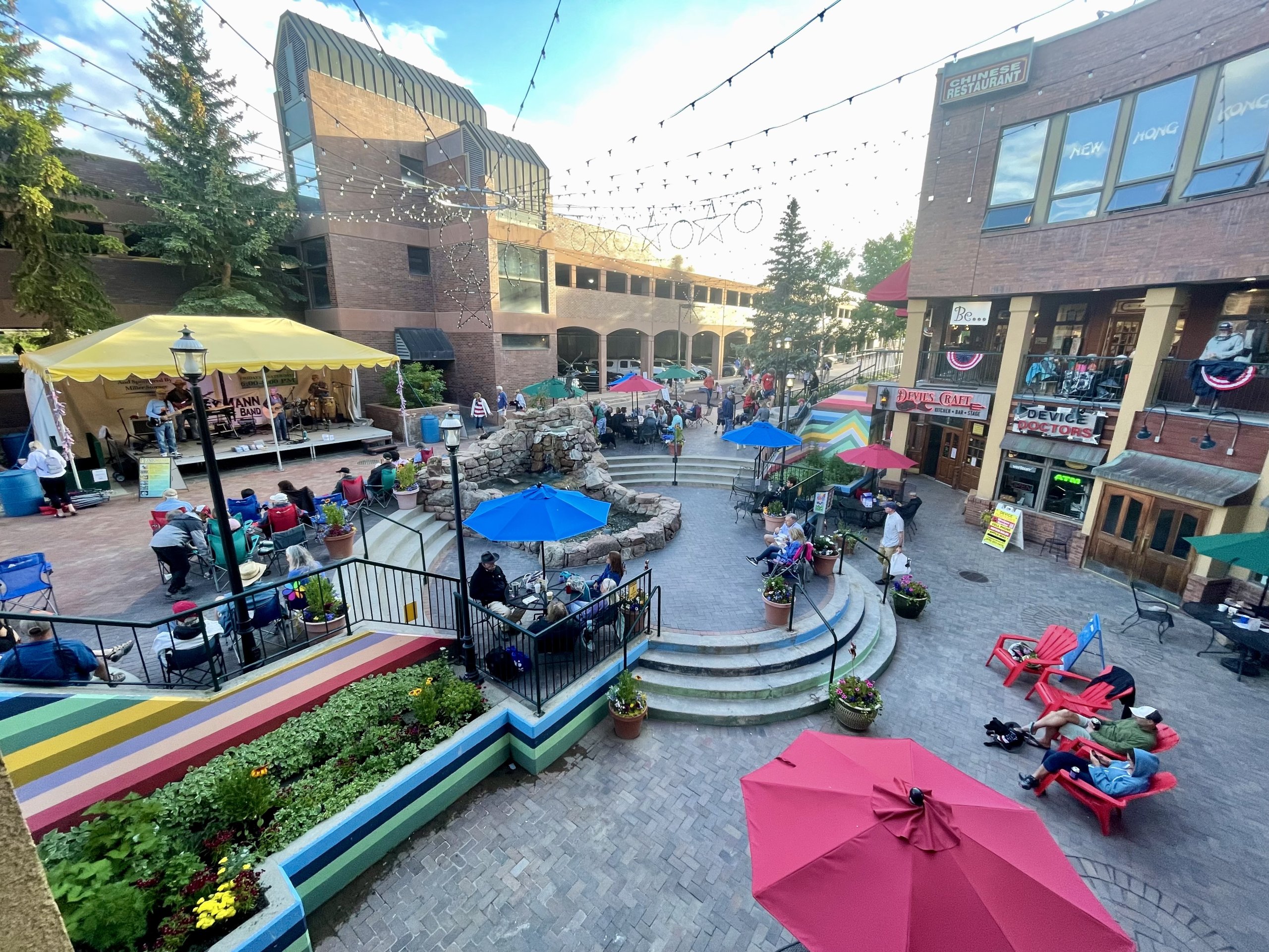 A vibrant town square bustling with people enjoying outdoor cafes, live music, and colorful gardens under a clear blue sky in Grand County Colorado.