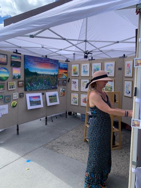 A woman in a floral dress and wide-brimmed hat stands in an outdoor art gallery tent, surrounded by colorful paintings and photographs in Grand County Colorado.