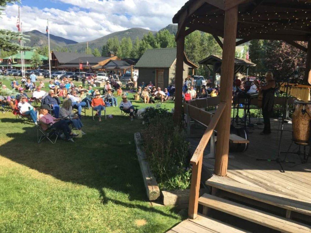 A lively outdoor concert in a grassy park with a wooden stage, surrounded by mountains and a clear blue sky in Grand County Colorado.