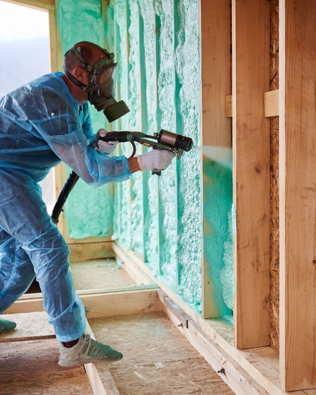 A construction worker in full protective gear sprays insulating foam onto wooden studs inside a building under construction in Grand County Colorado.
