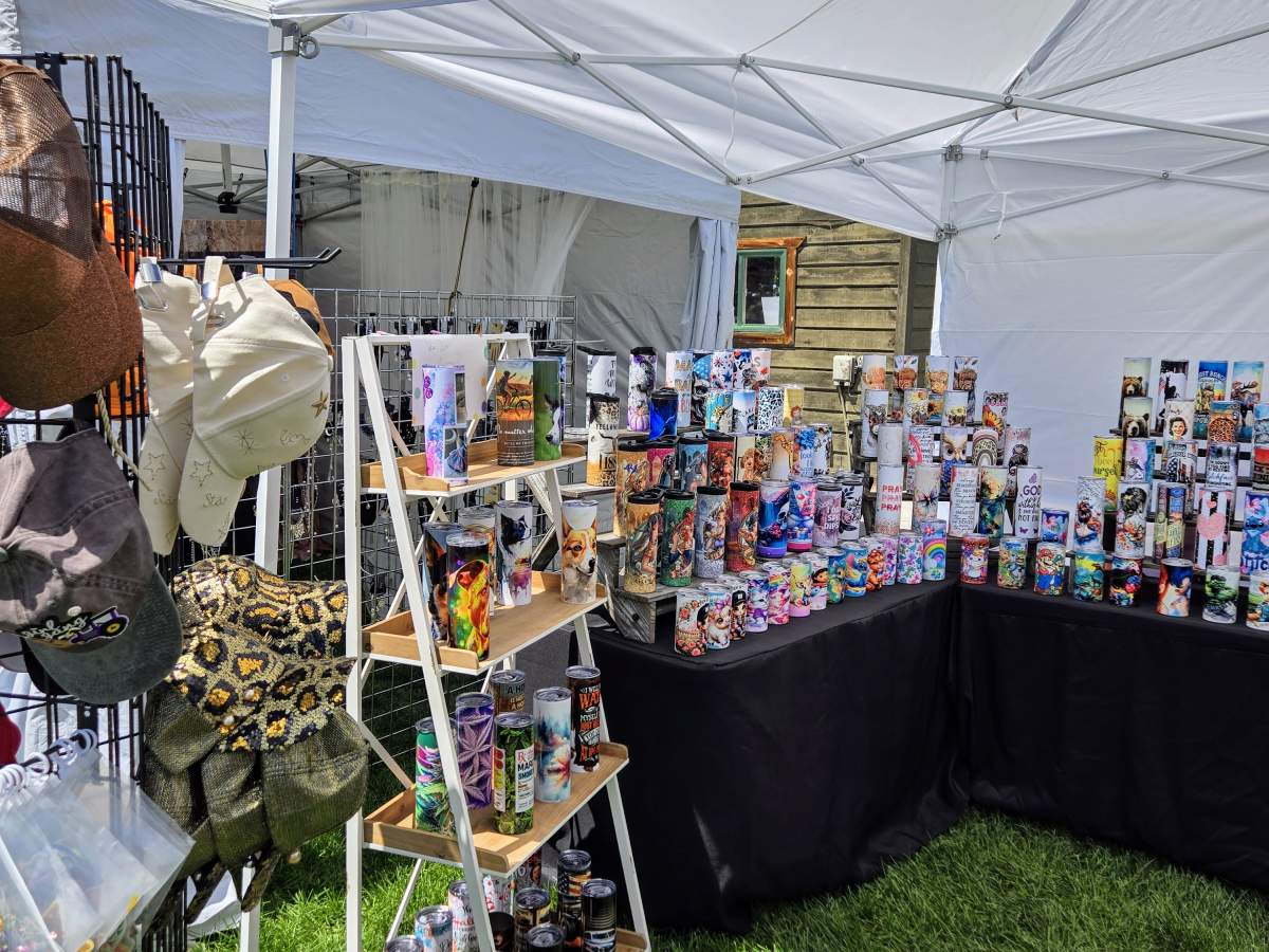 A vibrant outdoor market stall under a white tent, showcasing a colorful array of items including hats, bags, and custom-designed tumblers in Grand County Colorado.