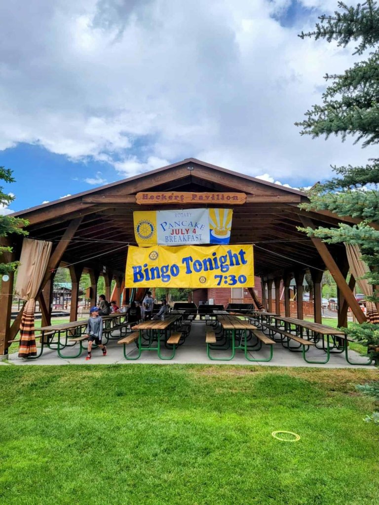 A large wooden pavilion with banners advertising a Rotary Pancake Breakfast and Bingo Tonight, surrounded by picnic tables and benches in a grassy area with trees in Grand County Colorado.