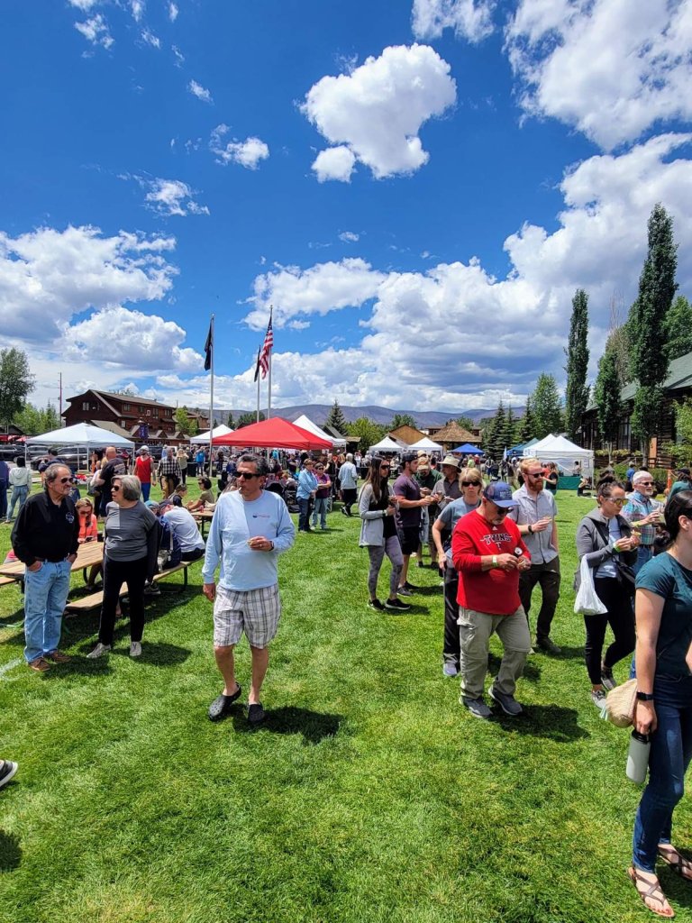 A lively outdoor festival with numerous attendees enjoying food, drinks, and activities under colorful tents on a sunny day in Grand County Colorado.
