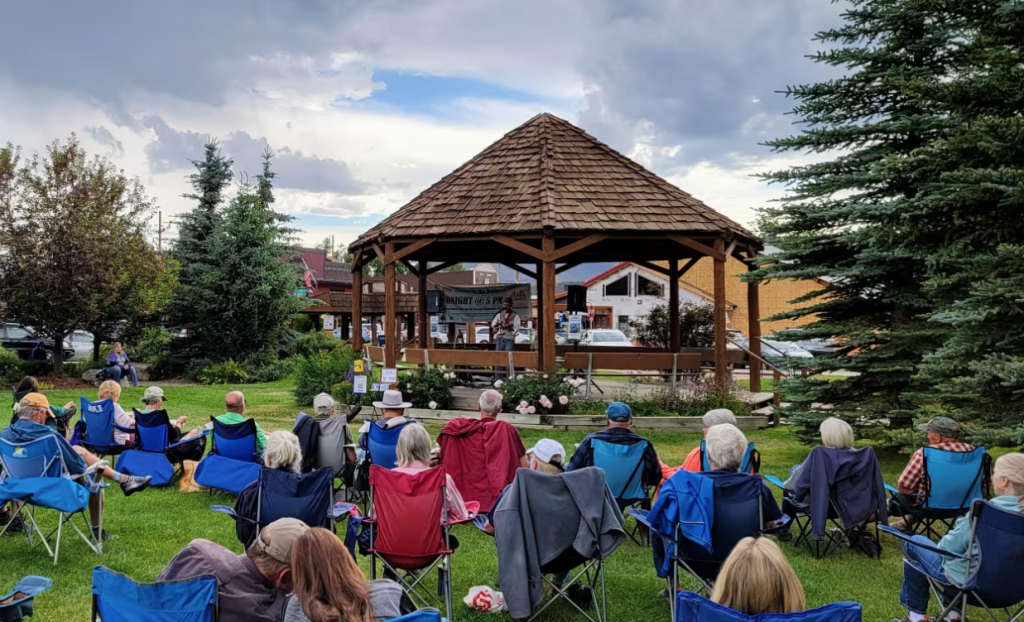 A group of people, mostly seniors, are seated on colorful folding chairs in a grassy area under a wooden gazebo, listening to a speaker at a small stage in Grand County Colorado.