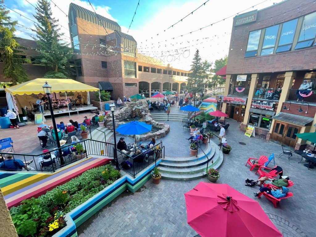 A vibrant outdoor plaza filled with people enjoying a sunny day, featuring colorful umbrellas, a stage with live music, and a variety of shops and restaurants in Grand County Colorado.
