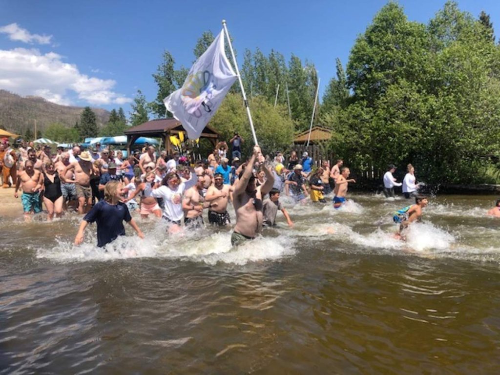 A large group of people joyfully running into a river, with one person waving a colorful flag, on a sunny day surrounded by lush greenery in Grand County Colorado.