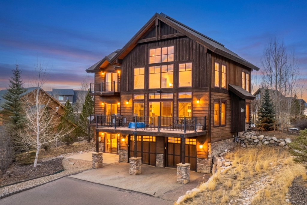 A modern, multi-story wooden house with large windows and balconies, nestled among trees with a mountain backdrop at dusk in Grand County Colorado.