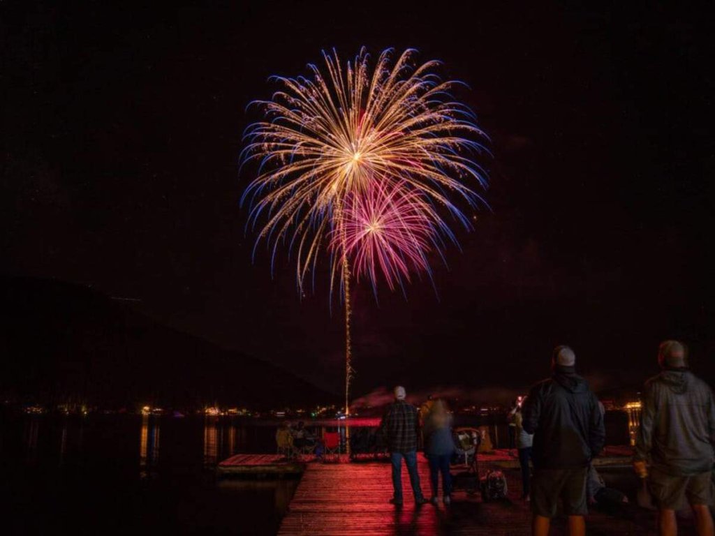 A vibrant fireworks display lights up the night sky over a serene lake, with people gathered on a wooden dock to watch the spectacle in Grand County Colorado.