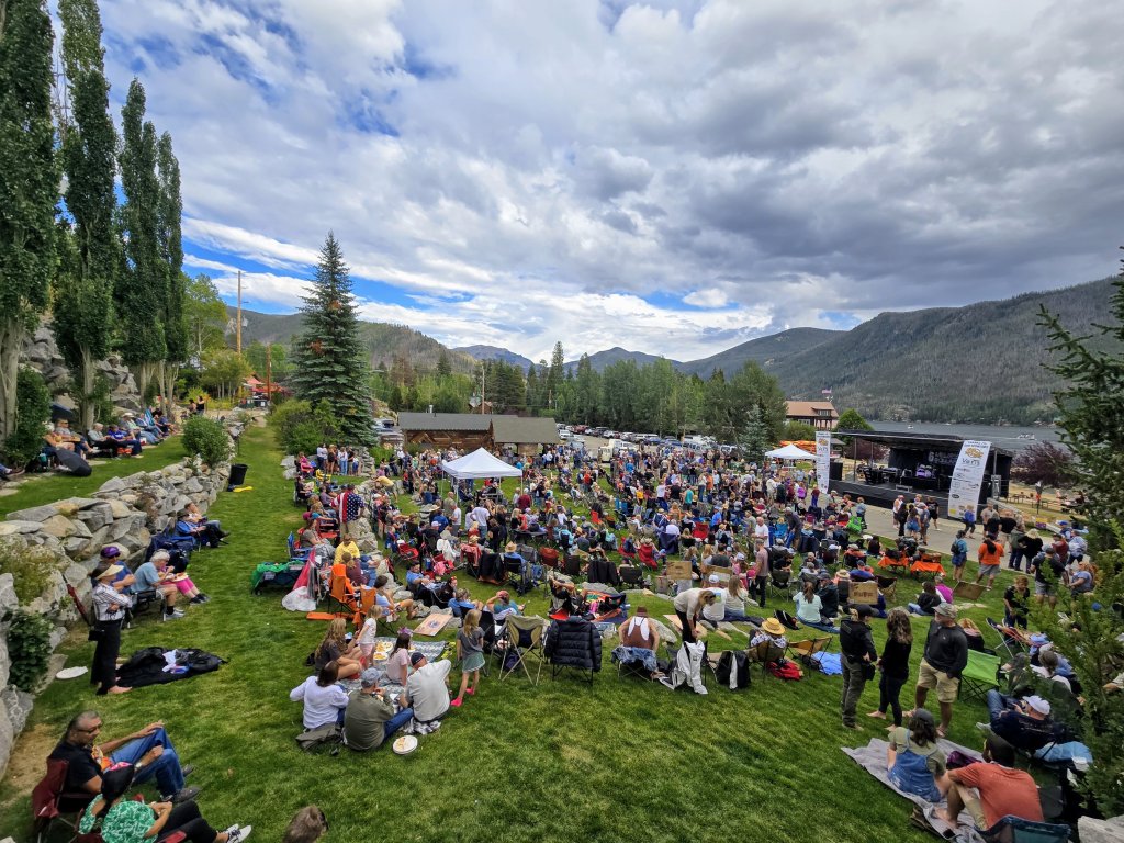 A lively outdoor festival with a large crowd gathered on a grassy field, surrounded by tall trees and mountains, with a stage set up for performances in Grand County Colorado.