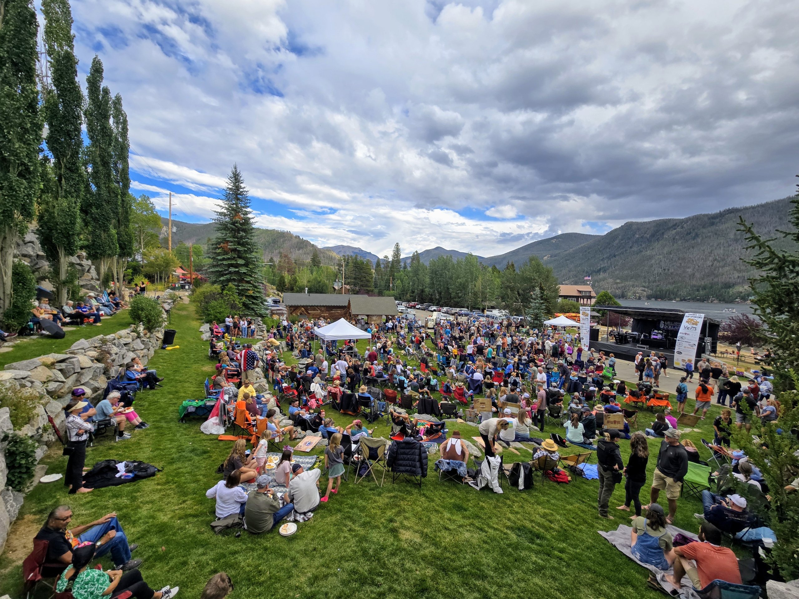 A lively outdoor festival with a large crowd gathered on a grassy field, surrounded by tall trees and mountains, with a stage set up for performances in Grand County Colorado.