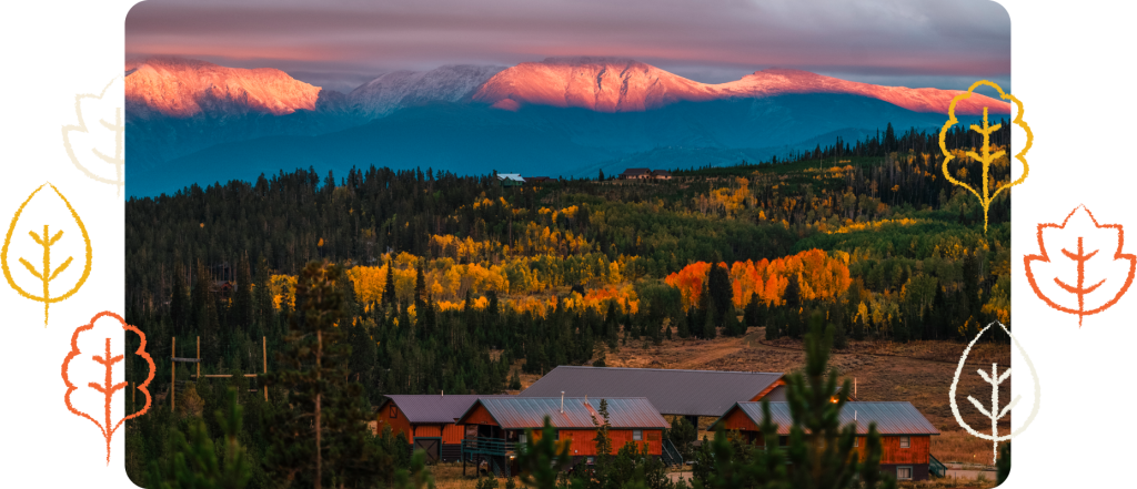 Houses sit among autumn trees with vibrant foliage, backed by forested hills and snow-capped mountains under a dramatic pink and purple sky.