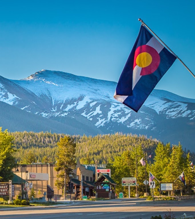 The Colorado state flag waves before a snow-capped mountain range and a small town in Grand County Colorado.
