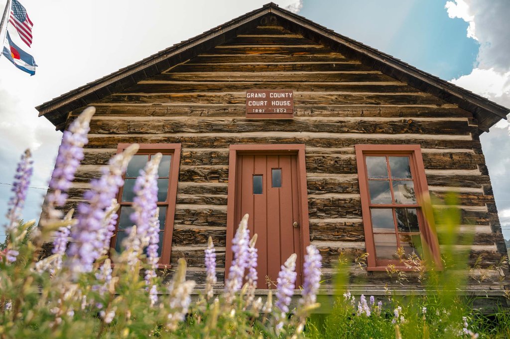 Historic Grand County Courthouse at Pioneer Village Museum, featuring a rustic log exterior, red-trimmed windows and wildflowers in the foreground.