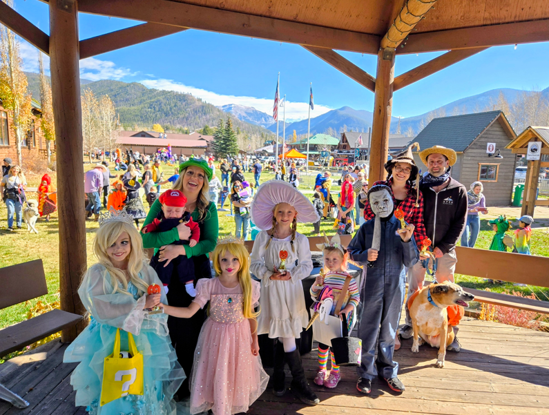 A group of children and adults in Halloween costumes posing under a wooden pavilion in Grand County Colorado.